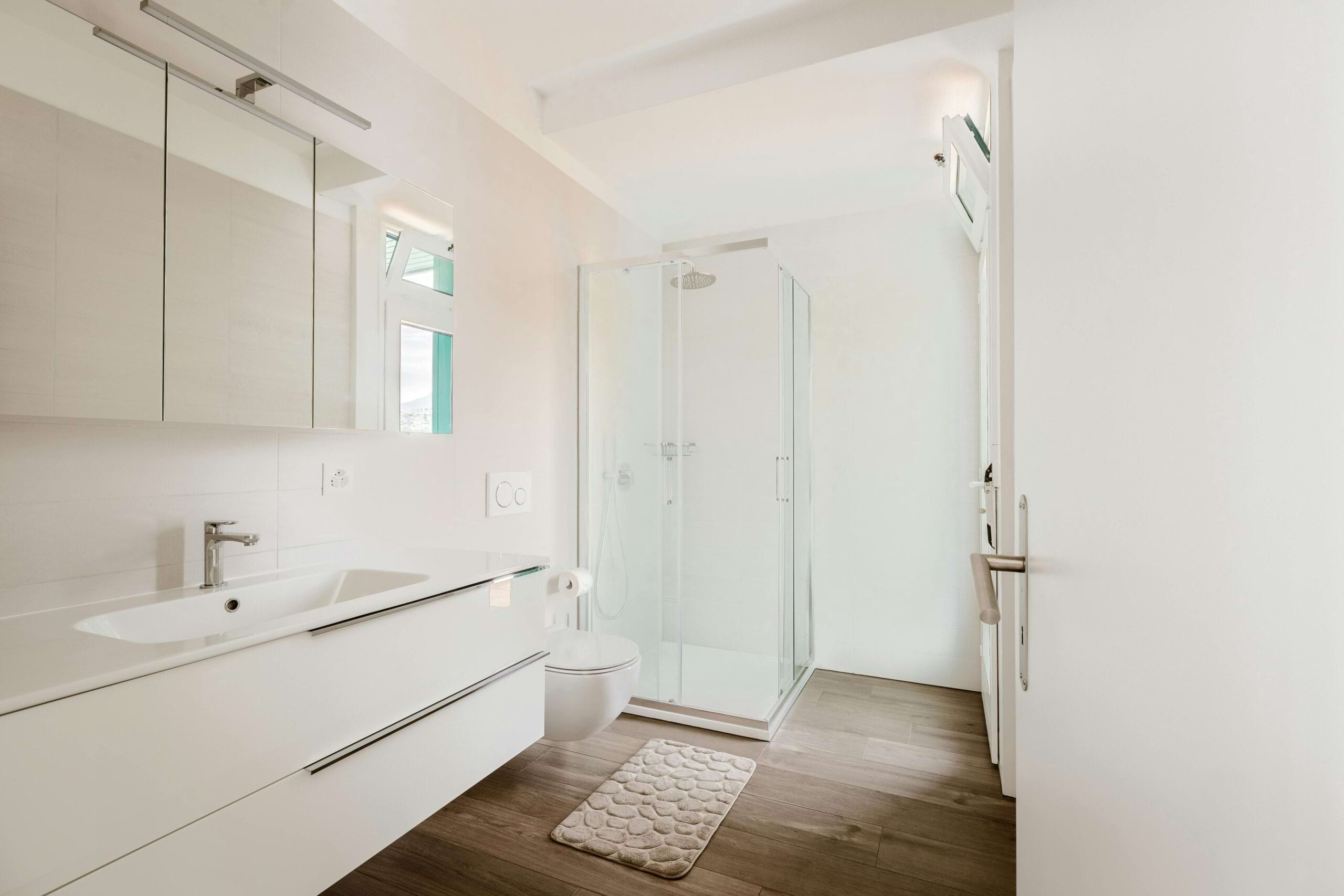 Helles Badezimmer mit Dusche, Waschbecken, Spiegel, WC und Holzfußboden. Ein Duschvorleger liegt am Boden.