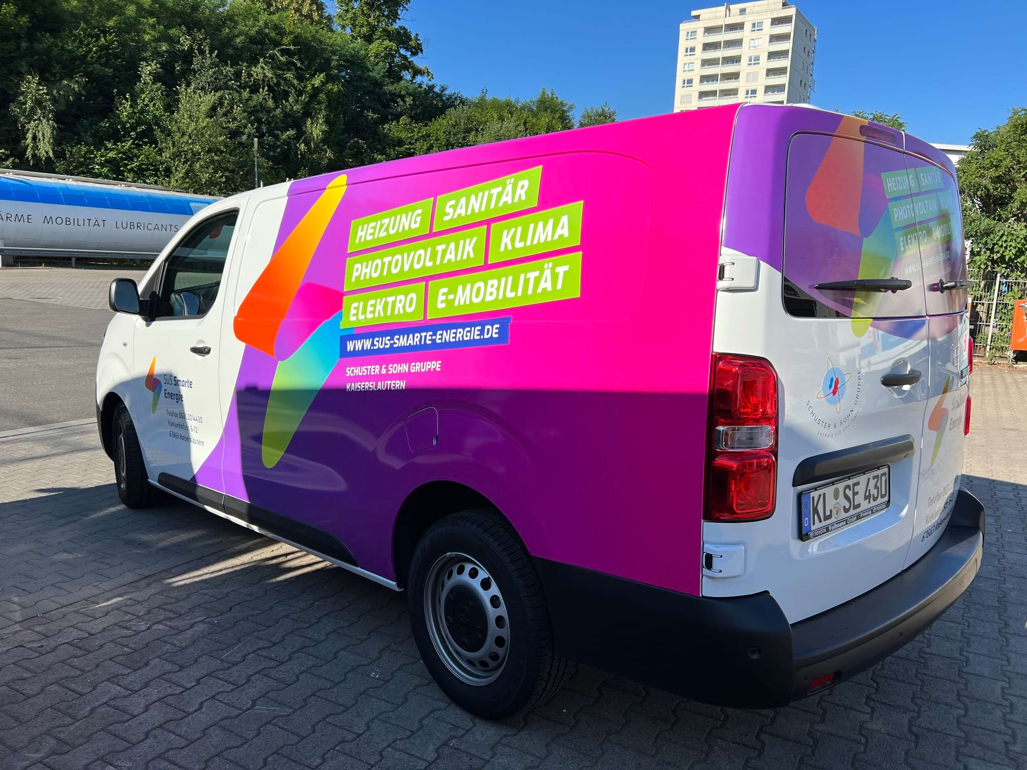 Ein weißer Van mit buntem Werbeaufdruck für Heizungs-, Sanitär- und Elektrodienstleistungen steht auf einem Parkplatz.