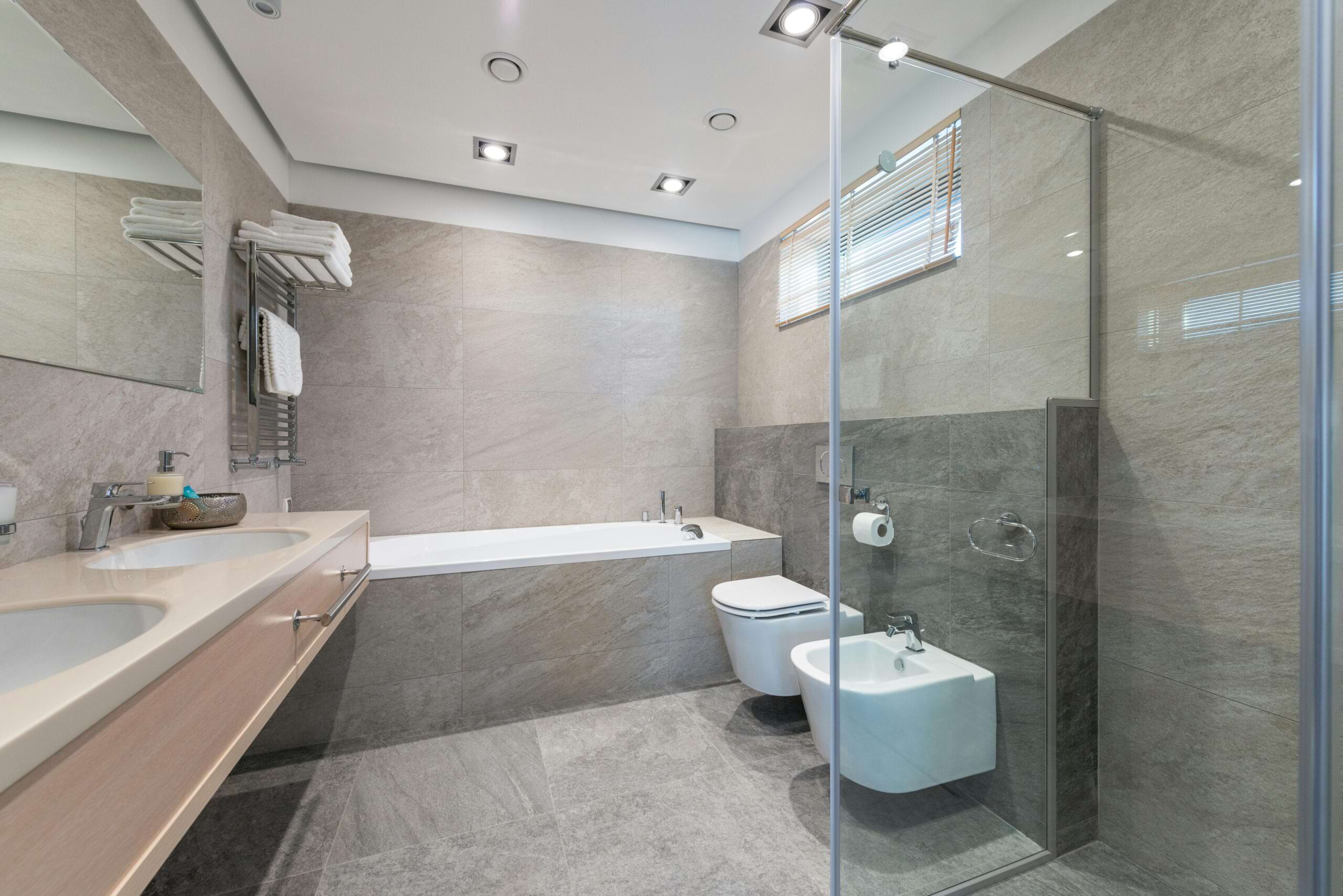 Geräumiges Badezimmer mit Badewanne, WC, Waschbecken, Dusche und handtuchhaltendem Regal. Graue Fliesen an Wänden und Boden.