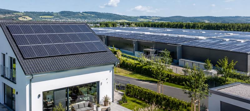 Ein Wohnhaus mit Solaranlagen auf dem Dach steht vor einer großen, bepflanzten Fläche und modernen Gebäuden, umgeben von Berg