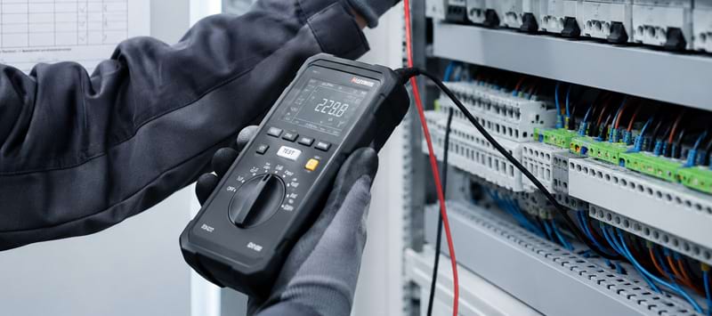 Ein Handwerker hält ein Multimeter, während er Kabel in einem Schaltkasten überprüft. Elemente der elektrischen Verkabelung s